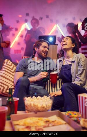 Junge Frau lacht und spricht mit dem jungen Mann, während sie auf dem Sofa sitzen und auf der Party Bier trinken und Popcorn essen Stockfoto