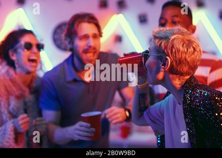 Junge Frau mit kurzen blonden Haaren, die Bier trinkt, und ihre Freunde, die sie unterstützen, haben Spaß auf der Party Stockfoto