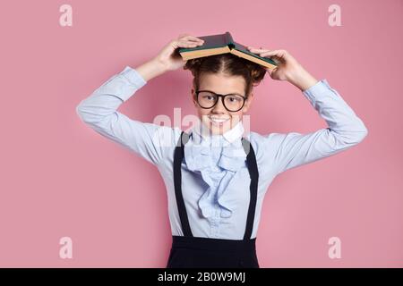 Lustige kleine Mädchen in Brille und Schuluniform mit Buch Über Kopf Stockfoto