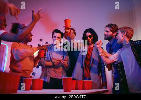 Junge Frau in einer Sonnenbrille, die das Glas Bier großzog, und sie wird es mit ihren Freunden trinken, die in ihrer Nähe auf der Party stehen Stockfoto
