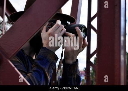 Man Fotograf, der ein Foto durch eine große rote Metall-Brücke mit einem braunen Cowboyhut und einem Square-Hemd nimmt, moderne Menschen Stockfoto