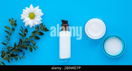 Set aus Kosmetikflaschen, Gesichtscreme und Lotion auf blauem Hintergrund mit grünem Ast und Daisy. Glasbecher und Spray mit weißer Kamille, Hautkanne Stockfoto
