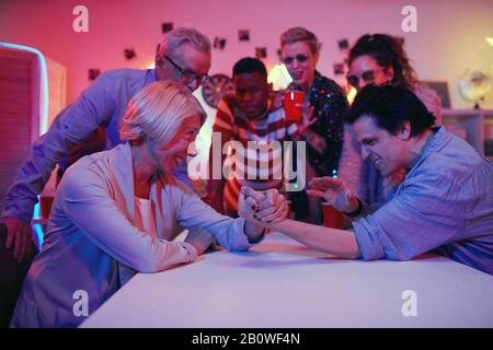 Die Leute, die am Tisch sitzen, halten die Hände und rivalisieren miteinander, während ihre Freunde sie auf der Party unterstützen Stockfoto