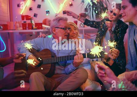 Liebevolles Senioren-Paar, das auf Gitarre spielt und den guten Abend mit ihren Freunden zu Hause auf Party genießt Stockfoto