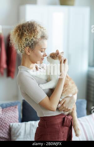 Junge Frau mit lockigem Haar, die steht und ihre niedliche Katze umarmt, die sie sehr liebt Stockfoto