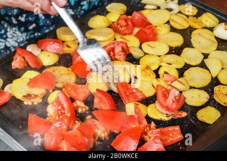 Kartoffeln und Tomaten auf kleine Stücke geschnitten, die auf dem elektrischen Grill gegrillt werden, verwischte Hand, die die Stahlgabel hält, die Raucherstücke über heiße Oberfläche bewegt Stockfoto
