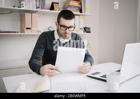 Freiberufler beschäftigt sich mit Papierkram im feigen Raum. Mann, der eine Brille mit schwarzem Rahmen trägt, der in Papierblätter im feigen Raum blickt. Stockfoto