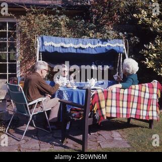 1960er Jahre, historisch, Frühjahrszeit und drei ältere Menschen draußen auf einer Terrasse sitzen auf den Gartenmöbeln der Ära, eine Schaukel, Bank und Klappstuhl, eine Tasse Tee, England, Großbritannien. Stockfoto