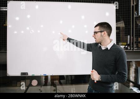 Junger Dozent, der vor dem Whiteboard steht. Lehrer zeigt auf leere weiße Tafel und hält Marker im Hörsaal. Stockfoto