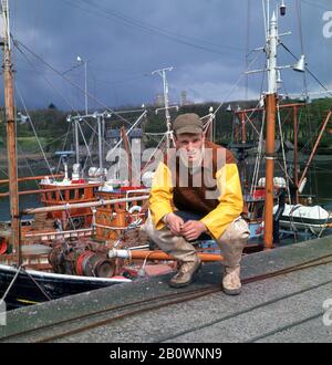 1960er Jahre, historisch, ein junger schottischer Fischer in seiner wasserdichten Angelkleidung und Kappe, kniend für ein Foto am Hafen von Stornoway, Isle of Lewis, Scottish Highland, Schottland, Großbritannien. Stockfoto