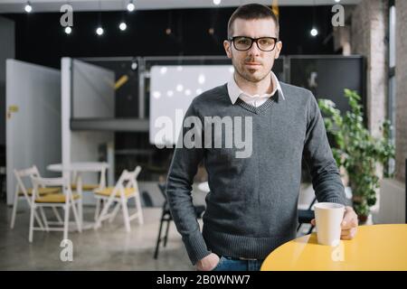 Mitarbeiter, der sich mit einer Tasse Kaffee im Hörsaal ausruht. Bärtiger Arbeiter mit Brille und formeller Kleidung, die eine Kaffeetasse neben dem gelben Tisch hält. Stockfoto