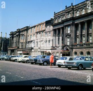 Ende der 1960er Jahre, historisch, eine Dame in einem orangen Kleid, die neben Autos der Ära stand, parkte vor dem George Hotel, George Street, Edinburgh, Schottland, Großbritannien, mit Autos der Ära, die draußen geparkt waren. Stockfoto