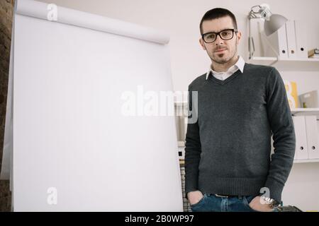 Business Trainer steht neben dem Flipchart Board. Junger Geschäftsmann, der in der Nähe eines Flipcharts mit Händen in den Taschen steht und in die Kamera blickt. Stockfoto