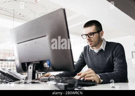 Konzentrierter Mann, der im Büro an einem Computer arbeitet. Bärtiger Mitarbeiter, der Computer verwendet und auf einem Büroschreibtisch mit Desktop sitzt. Stockfoto