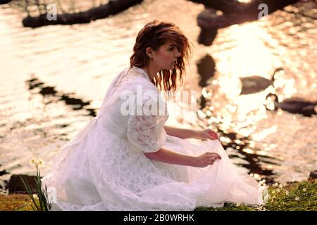 Junge Frau in weißem Kleid sitzt am See Stockfoto