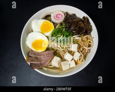 Ramen: Asiatische Nudelsuppe mit Rindfleisch, Eiern, Pilzen, Gemüse, Tofu, Algen und Naruto in einer Schüssel. Schwarzer Hintergrund. Draufsicht. Stockfoto