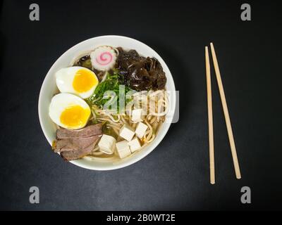 Ramen: Asiatische Nudelsuppe mit Rind, Eiern, Pilzen, Gemüse, Tofu, Algen und Naruto in einer Schüssel mit Essstäbchen auf der Seite. Schwarzer Hintergrund. Top vie Stockfoto