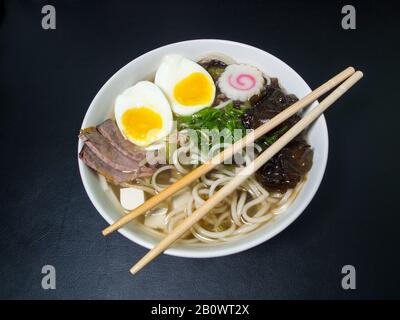 Ramen: Asiatische Nudelsuppe mit Rindfleisch, Eiern, Pilzen, Gemüse, Tofu, Algen und Naruto mit Essstäbchen auf der Schüssel. Schwarzer Hintergrund. Draufsicht. Stockfoto