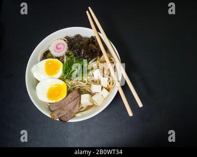 Ramen: Asiatische Nudelsuppe mit Rindfleisch, Eiern, Pilzen, Gemüse, Tofu, Algen und Naruto in einer Schüssel mit Essstäbchen. Schwarzer Hintergrund. Draufsicht. Stockfoto