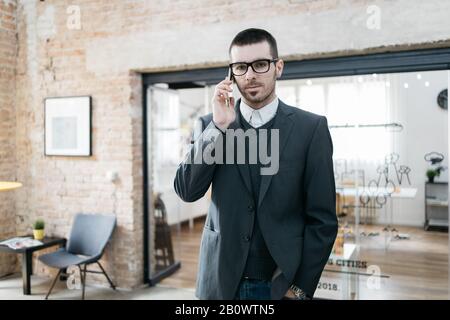 Porträt des ernsthaften Kerls mit gefalteten Händen. Junger Geschäftsmann, der Brille mit gefalteten Armen trägt, die im Büro stehen. Stockfoto