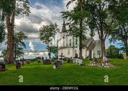 Weiße Holzkirche umgeben von Grün, Friedhof mit Gräbern auf dem Boden in der grünen Wiese begraben. Steinkjer, Kreis Trøndelag, Norwegen Stockfoto