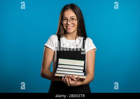 Asiatisches Studenten-Mädchen auf blauem Hintergrund im Studio hält Stapel von Universitätsbüchern aus der Bibliothek. Frau lächelt, sie ist glücklich, einen Abschluss zu machen. Stockfoto