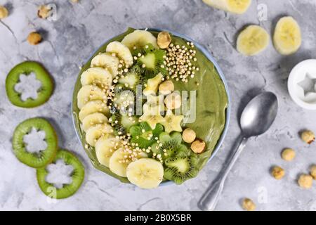 Grüne Fruchtkale-Smoothie-Schüssel mit sternförmigen Bananen, Kiwi, Haselnuss und gebucktem Quinoa-Korn auf grauem Grund Stockfoto