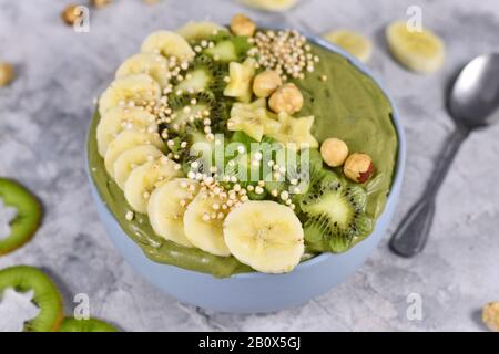 Grüne Fruchtkale-Smoothie-Schüssel mit sternförmigen Bananen, Kiwi, Haselnuss und gebucktem Quinoa-Korn auf grauem Grund Stockfoto