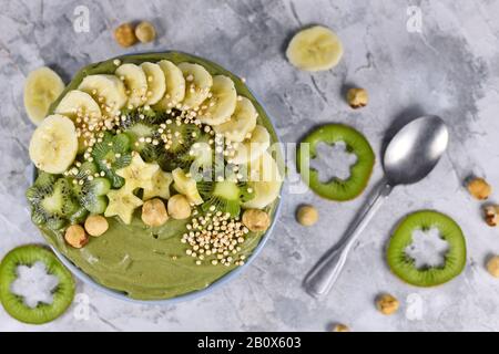 Grüne Fruchtkale-Smoothie-Schüssel mit sternförmigen Bananen, Kiwi, Haselnuss und gebucktem Quinoa-Korn auf grauem Grund Stockfoto