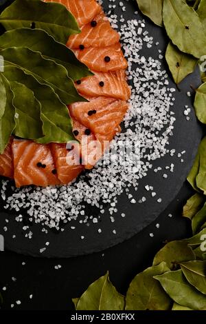 Frisch gesalzene rote Fische, die in Scheiben geschnitten werden, mit grobem Salzachenblatt und schwarzen Pfeffererbsen auf einem Schiefersubstrat. Stockfoto