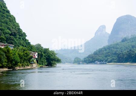 Der Fluss Li in Yangshuo, Guilin, China, mit einem Haus, entfernten Gebäuden und Karstbergen, die sich im Nebel am Horizont verstecken Stockfoto