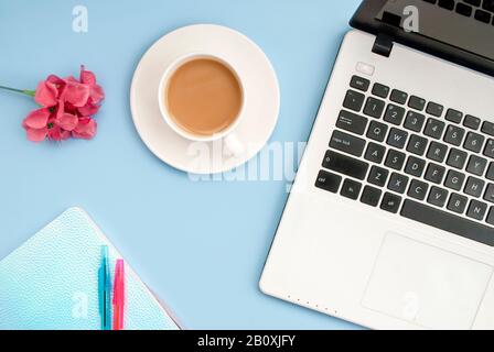 Weiße Tasse Kaffee-Notizblock, pinke Blume, Stifte, Laptop-Tastatur auf blauem Hintergrund. Flaches Layout. Kopierbereich. Draufsicht. Stockfoto