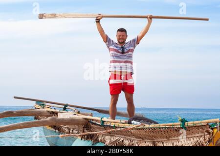 Porträt eines Mannes mit mittlerem Bart im Hemd und roten Shorts auf einem traditionellen Fischerboot mit einem Paddel in den Händen. Paddel hält ausgestreckt Stockfoto