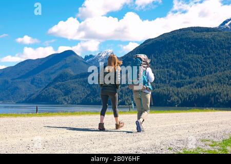 Zwei junge Frauen gehen auf Schotterstraße neben einem See mit Bergen im Hintergrund. Stock Foto. Stockfoto