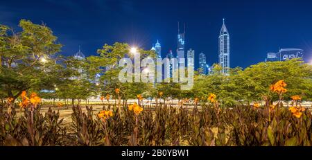 Grünfläche mit Palmen und Blumen an einer Autobahnanschlussstelle der Scheich Zayed Road mit Blick auf Wolkenkratzer in Marina bei Nacht, New Dubai, Vereinigte Arabische Emirate, Stockfoto