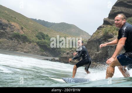 Zwei männliche Surfer in schwarzen Wetsuits reiten auf einer grünen Bruchwelle am Piha Beach, Auckland, Neuseeland. Felsige Küste mit blauem Himmel Sommertag. Stockfoto