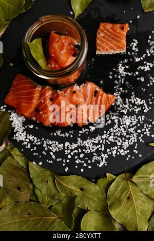Zutaten zum Salzen von rotem Fisch mit Lorbeerblatt und schwarzem Pfeffer in einem Glasbecher auf einem Schiefersubstrat. Stockfoto