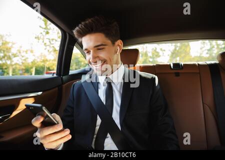 Bild eines jungen kaukasischen erfolgreichen Geschäftsmannes in formellem schwarzen Anzug mit Ohrstöpseln und Handy während der Fahrt im Auto Stockfoto