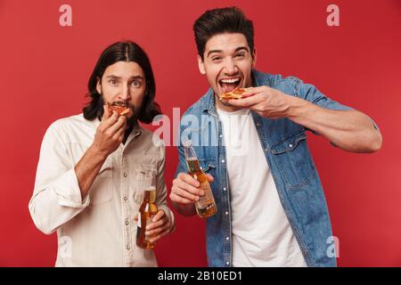 Foto von jungen aufgeregten Jungs, die Bier trinken und Pizza essen, isoliert über rotem Hintergrund Stockfoto