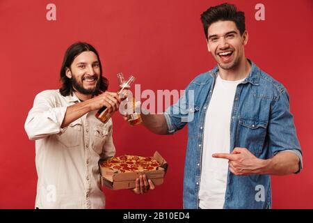 Foto von jungen lächelnden Jungs, die Bier trinken und Pizza essen, während sie den Finger isoliert über den roten Hintergrund zeigen Stockfoto