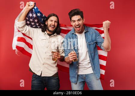 Foto von jungen begeisterten Jungs Fans, die Biere und amerikanische Flagge halten, während sie das Sportspiel beobachten, das über roten Hintergrund isoliert ist Stockfoto