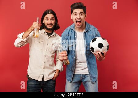 Foto von begeisterten Jungs Fans, die Biere und Fußball halten, während sie Sportspiele über rotem Hintergrund beobachten Stockfoto