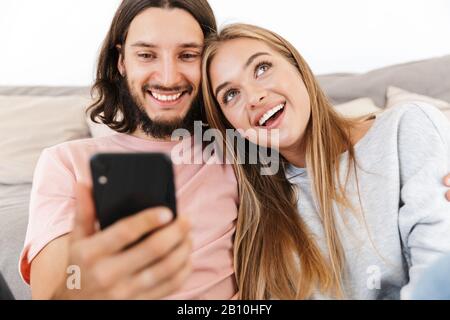 Bild eines lächelnden träumenden jungen Liebespaares auf dem Sofa zuhause mit dem Handy. Stockfoto