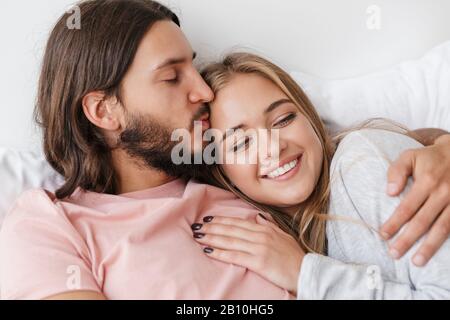 Wunderschönes, lächelndes junges Paar, das zu Hause im Bett liegt und sich umarmt Stockfoto