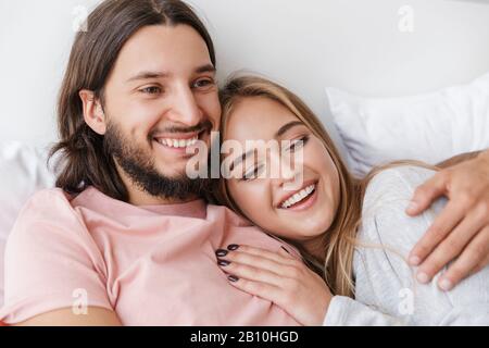 Wunderschönes, lächelndes junges Paar, das zu Hause im Bett liegt und sich umarmt Stockfoto