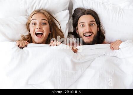 Draufsicht über ein wunderschönes, aufgeregtes junges Paar, das in einer Decke bedeckt ist, die zu Hause im Bett liegt, mit Blick auf die Kamera Stockfoto