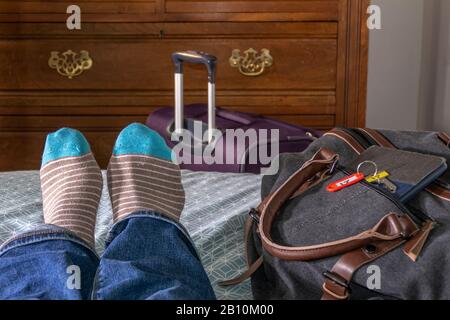 Nahe POV von einem Mann, der Socken trägt, auf einem Bett in einem Hotelzimmer/Motel liegend, gerade eingecheckt, mit Gepäck und Zimmerschlüssel erschossen. Stockfoto