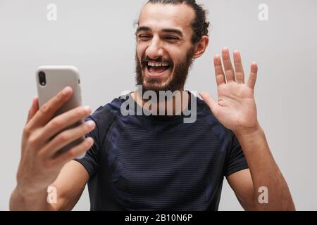 Bild eines bärtigen fröhlichen Sportlers, der einen Trainingsanzug trägt und dabei auf einem Smartphone spricht, das über weißen Hintergrund isoliert ist Stockfoto