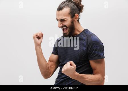 Bild des fröhlichen brünetten Sportlers mit einem Trainingsanzug, der schreiend und gestikulierend als Sieger über weißem Hintergrund isoliert ist Stockfoto