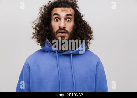 Bild eines eingeschüchterten, unrasierten Mannes in einem lässigen Sweatshirt, das Wunder an der Kamera ausdrückt, die über weißem Hintergrund isoliert ist Stockfoto